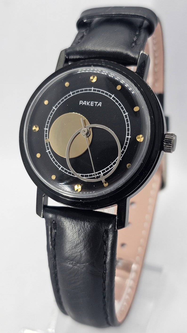 Puede incluir: Un reloj de pulsera negro con una correa de cuero negro. La esfera del reloj es negra con detalles dorados y presenta un dise&ntilde;o &uacute;nico de fase lunar. La esfera del reloj tambi&eacute;n tiene la palabra "PAKETA" impresa en ella.