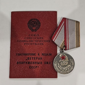 May include: A red booklet with the text "СОЮЗ СОВЕТСКИХ СОЦИАЛИСТИЧЕСКИХ РЕСПУБЛИК" and "УДОСТОВЕРЕНИЕ К МЕДАЛИ "ВETERAN" ВООРУЖЕННЫХ СИЛ СССР"  is next to a silver medal with a red star and the text "ВETERAN ВООРУЖЕННЫХ СИЛ СССР".