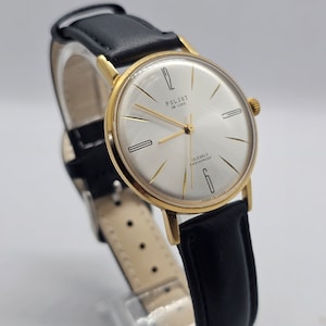 May include: A vintage gold-toned Poljot De Luxe wristwatch with a silver face and black leather band. The watch face features gold hands and markers, with the text "POLJOT DE LUXE" and "13 JEWELS SHOCKPROOF". The watch is marked "MADE IN USSR".