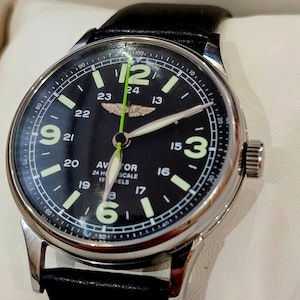 Könnte beinhalten: Ein schwarzes Zifferblatt mit weißen und grünen Zeigern und einem grünen Sekundenzeiger. Das Zifferblatt hat eine 24-Stunden-Skala und den Text "Aviator 24 Hour Scale 19 Jewels".