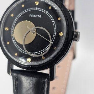 Puede incluir: Un reloj de pulsera negro con una correa de cuero negro. La esfera del reloj es negra con un c&iacute;rculo dorado y un c&iacute;rculo blanco con una aguja plateada. La esfera del reloj tambi&eacute;n tiene puntos dorados alrededor del borde y la palabra "PAKETA" impresa en blanco.