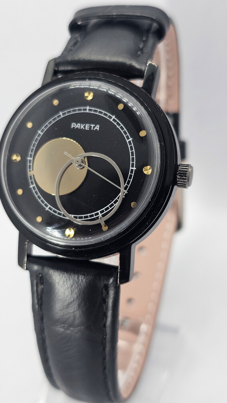 Puede incluir: Un reloj de pulsera negro con una correa de cuero negro. La esfera del reloj es negra con un c&iacute;rculo dorado y un c&iacute;rculo blanco con una aguja plateada. La esfera del reloj tambi&eacute;n tiene puntos dorados alrededor del borde y la palabra "PAKETA" impresa en blanco.