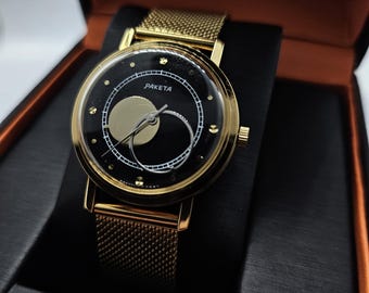 Op de afbeelding: Een goudkleurig Paketa horloge met een zwarte wijzerplaat en gouden accenten. Het horloge heeft een mesh band en wordt gepresenteerd in een zwarte doos met een oranje interieur. De wijzerplaat heeft een uniek ontwerp met een cirkelvormig patroon en de merknaam "Paketa".