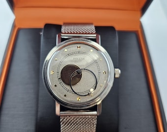 Puede incluir: Un reloj de pulsera plateado con una correa de malla plateada. La esfera del reloj es plateada con una superficie texturizada y presenta un círculo negro con un círculo negro más pequeño en su interior. La esfera del reloj también tiene detalles dorados y la palabra "PAKEta" impresa en ella.