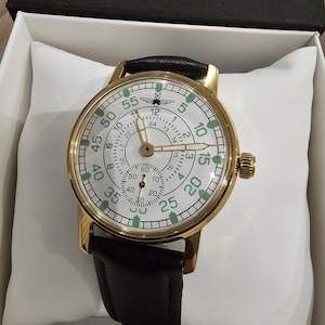 May include: A gold-toned wristwatch with a white face, black leather band, and a second dial. The watch face features black and green numerals and markings. The watch is presented in a white box.