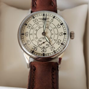 Op de afbeelding: Een vintage horloge met een crèmekleurige wijzerplaat, zwarte cijfers en een kleine secondewijzerplaat. Het horloge heeft een zilverkleurige kast en een bruine leren band. De wijzerplaat toont de cijfers van 1 tot 60, en de wijzers zijn zwart.