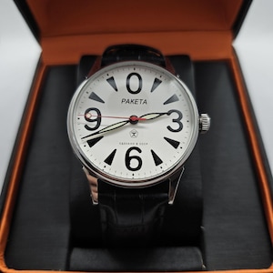 Peut inclure: Une montre argentée avec un cadran blanc, affichant de grands chiffres noirs et la marque "PAKETA". La montre a un bracelet en cuir noir et est présentée dans un coffret noir et orange.