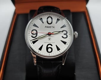 May include: A silver wristwatch with a white face, displaying large black numerals and the brand name "PAKETA". The watch has a black leather strap and is presented in a black and orange box.