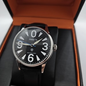 Könnte beinhalten: Eine schwarz-silberne Armbanduhr mit einem schwarzen Lederarmband. Das Zifferblatt der Uhr hat weiße Zahlen und einen blauen Sekundenzeiger. Das Zifferblatt der Uhr hat auch den Text "PAKEta" und "СДЕЛАНО В СССР".