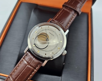 Puede incluir: Un reloj de pulsera plateado con una correa de cuero marrón. La esfera del reloj es blanca con marcas negras y la palabra "PAKETA" impresa en ella. El reloj tiene una complicación de fase lunar única.
