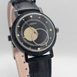 Puede incluir: Un reloj de pulsera negro con una correa de cuero negro y una esfera dorada. La esfera tiene un c&iacute;rculo dorado en el centro y la palabra "PAKETA" impresa en letras doradas.