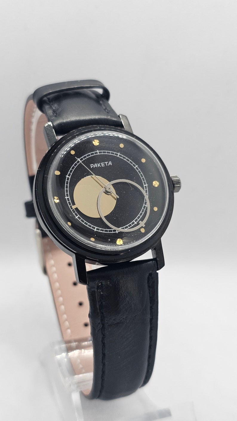 Puede incluir: Un reloj de pulsera negro con una correa de cuero negro y una esfera dorada. La esfera tiene un c&iacute;rculo dorado en el centro y la palabra "PAKETA" impresa en letras doradas.