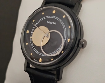 May include: A black wristwatch with a black leather band. The watch face has a black background with a gold circle and a smaller black circle inside. The watch face also has gold markings and the word "PAKETA" printed in gold.