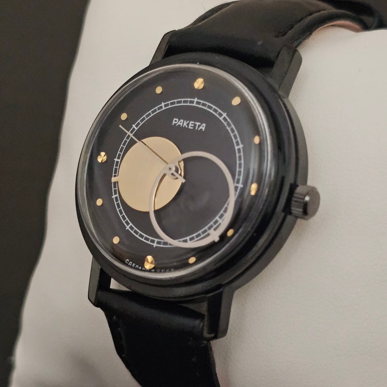 Puede incluir: Un reloj de pulsera negro con una correa de cuero negro. La esfera del reloj tiene un fondo negro con un c&iacute;rculo dorado y un c&iacute;rculo negro m&aacute;s peque&ntilde;o en su interior. La esfera del reloj tambi&eacute;n tiene marcas doradas y la palabra "PAKETA" impresa en oro.