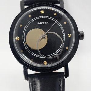 Puede incluir: Un reloj de pulsera negro con una correa de cuero negro. La esfera del reloj es negra con marcas blancas y un c&iacute;rculo dorado en el centro. La esfera del reloj tambi&eacute;n tiene el texto "РАКЕТА" en letras doradas.
