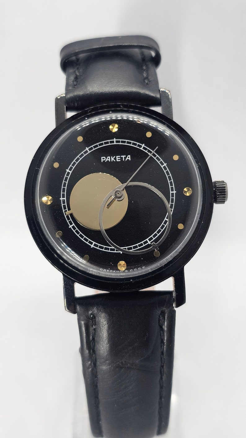 Puede incluir: Un reloj de pulsera negro con una correa de cuero negro. La esfera del reloj es negra con marcas blancas y un c&iacute;rculo dorado en el centro. La esfera del reloj tambi&eacute;n tiene el texto "РАКЕТА" en letras doradas.