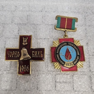 Könnte beinhalten: Zwei Vintage-Medaillen. Eine ist eine kreuzförmige Medaille mit einer Glocke und dem Text "ЧОРНО БИЛЬ 1986". Die andere ist eine sternförmige Medaille mit einem Band und dem Text "УЧАСТНИК ЛИКВИДАЦИИ ПОСЛЕДСТВИЙ АВАРИИ ЧАЭС". Beide Medaillen haben rote und goldene Akzente.