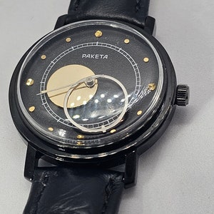 Puede incluir: Un reloj de pulsera negro con una correa de cuero negro. La esfera del reloj es negra con detalles dorados y la palabra "PAKEta" impresa en ella. El reloj tiene un dise&ntilde;o &uacute;nico de fase lunar.