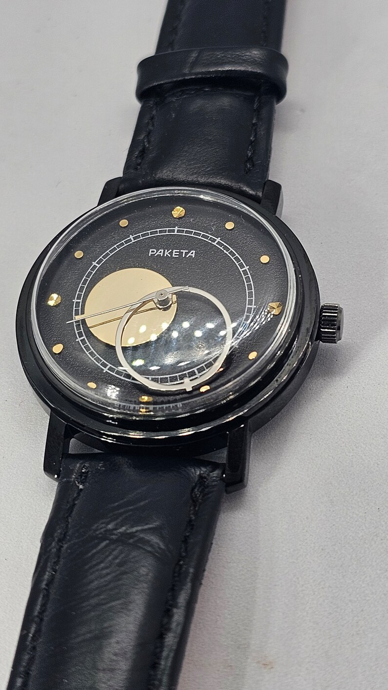 Puede incluir: Un reloj de pulsera negro con una correa de cuero negro. La esfera del reloj es negra con detalles dorados y la palabra "PAKEta" impresa en ella. El reloj tiene un dise&ntilde;o &uacute;nico de fase lunar.