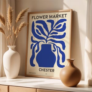 Può includere: Una stampa incorniciata con un design minimalista che presenta un vaso blu con una pianta stilizzata davanti a uno sfondo crema. Il testo "FLOWER MARKET" e "CHESTER" è stampato sulla stampa.