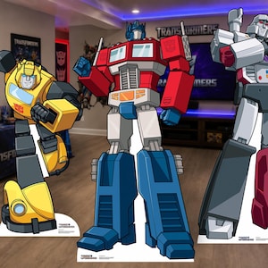 May include: Three life-sized cardboard standees featuring Transformers characters. Bumblebee is yellow and black, Optimus Prime is red, blue, and silver, and Megatron is white, grey, and maroon. Each figure displays the iconic Autobot or Decepticon logo.