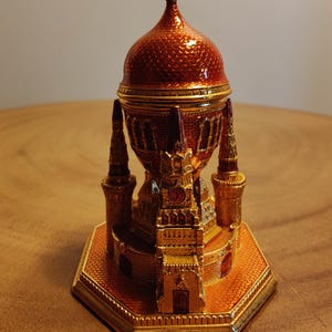 May include: An ornate, gold-coloured decorative object, possibly a church or cathedral. It has a red, textured dome and multiple spires. The hexagonal base and overall design are highly detailed and intricate, with a metallic finish.