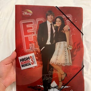 Może przedstawiać: Czerwona plastikowa teczka z grafiką High School Musical 3 Senior Year i logo Disney. Okładka przedstawia zdjęcie dwóch osób w strojach wizytowych, napis High School Musical 3 Senior Year oraz odręczne podpisy.