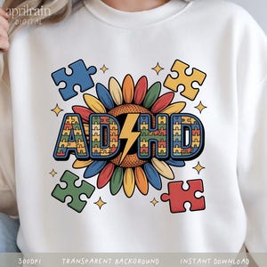 Puede incluir: Una sudadera blanca con un diseño gráfico de un girasol con pétalos multicolores. Las letras ADHD están escritas en una fuente negrita rellena con un patrón de piezas de rompecabezas, con un rayo amarillo reemplazando la letra H.
