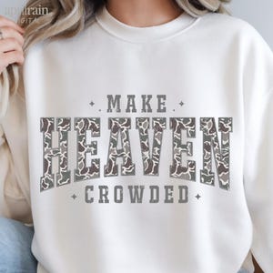 Puede incluir: Una sudadera blanca de cuello redondo con el texto MAKE HEAVEN CROWDED en fuente de bloque. La palabra HEAVEN está rellena con un patrón de camuflaje gris, blanco y gris oscuro, mientras que las palabras MAKE y CROWDED son de color gris sólido con pequeñas estrellas.