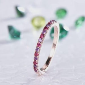 May include: A rose gold ring with a band of alternating red and purple gemstones. The ring is set against a blurred background with green and yellow accents, creating a sparkling effect. The ring is a delicate piece of jewelry.
