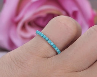 May include: A turquoise bead ring is shown on a finger. The ring features a band of small, round, light blue beads. The background is a soft pink, out-of-focus floral arrangement.