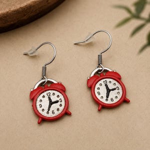 May include: A pair of red alarm clock earrings with silver-coloured hooks. The clock faces are white with black numbers and hands, and the time displayed is approximately 12:20. The earrings are designed to dangle.
