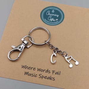 May include: Silver-colored keychain with a lobster clasp, a key ring, the letter "C", and a musical note charm. The keychain is on a brown card with the text "Where Words Fail Music Speaks."