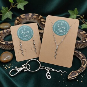 May include: A selection of snake-themed jewellery and accessories. Features a silver-toned necklace, earrings, and a keyring with a letter "G" charm. Each item is presented on a brown card with a "Charming Gifts" logo.
