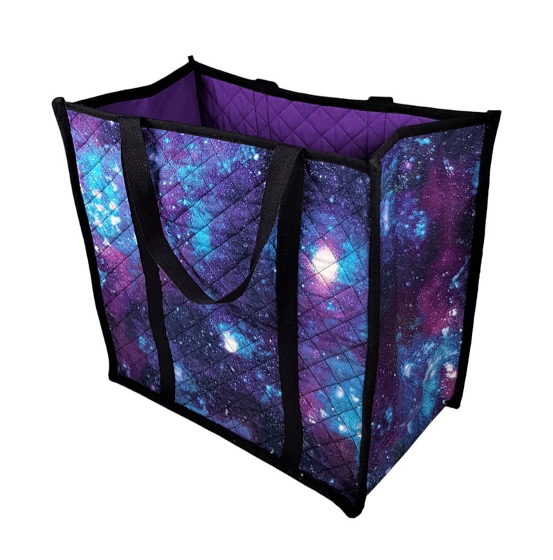 Puede incluir: Una bolsa de lona acolchada grande con asas negras y un estampado de galaxia en tonos morados, azules y negros.