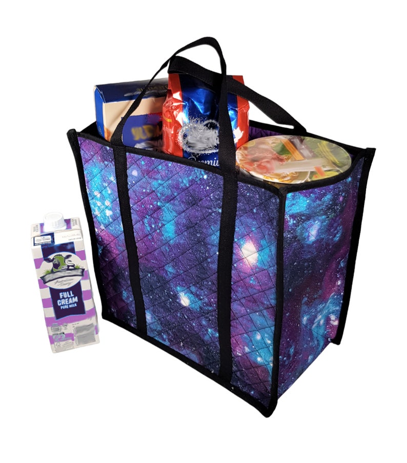 Puede incluir: Una bolsa de mano acolchada con estampado de galaxia en tonos morados, azules y negros. La bolsa tiene asas negras y est&aacute; llena de comestibles, incluyendo una caja de leche, una caja de cereales y un recipiente de fideos.