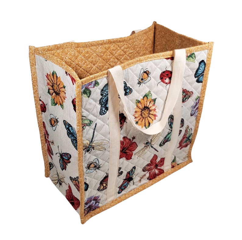 Puede incluir: Una bolsa de mano grande acolchada con fondo blanco y un estampado colorido de mariposas, flores y lib&eacute;lulas. La bolsa tiene dos asas blancas y un ribete acolchado marr&oacute;n.