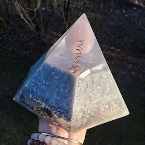Pode incluir: Um dispositivo de orgone transparente em forma de pirâmide com uma esfera de quartzo rosa e uma bobina de cobre no interior. A pirâmide tem uma base prateada e cinzenta. Segurado em uma mão com pulseiras de contas.