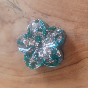May include: A green and silver flower-shaped resin ornament with gold flecks. The ornament is made of clear resin with green and silver glitter embedded in it.