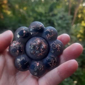 May include: A dark purple, flower-shaped object with copper-colored accents and embedded green and gold flecks. The object is held in a hand against a blurred green background.