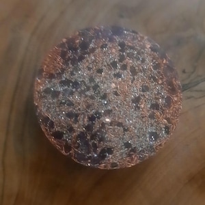 May include: A round, handcrafted object with a copper wire exterior and a dark, speckled interior. The copper wire is intricately woven, creating a textured surface. The interior appears to have small, dark stones or beads.