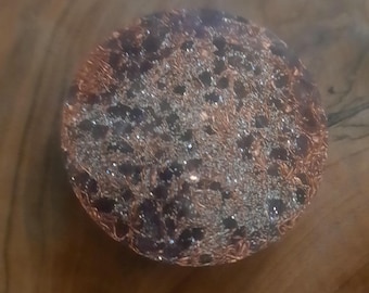 Può includere: Un oggetto rotondo e fatto a mano con un esterno in filo di rame e un interno scuro e maculato. Il filo di rame è intrecciato in modo intricato, creando una superficie strutturata. L'interno sembra avere piccole pietre o perle scure.