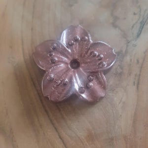 May include: A rose gold metal flower charm with a five-petal design and small raised dots around the center.