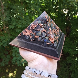 Könnte beinhalten: Ein pyramidenförmiges Objekt aus Kunstharz mit eingebetteten dunkelgrauen und blauen Steinfragmenten, die mit Kupferspänen vermischt sind. Die Basis der Pyramide besteht aus einer soliden schwarzen Schicht, während der obere Teil durchscheinend ist.