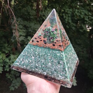May include: A clear resin pyramid containing a green and pink zoisite stone at the top, surrounded by a layer of copper wire shavings and small black stones. The base section is filled with green glass fragments and dark brown material.