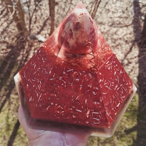 Puede incluir: Una pirámide de resina roja y transparente con una bobina de cobre y una inclusión esférica. La pirámide se sostiene en una mano, con un fondo natural al aire libre. Es un objeto decorativo.