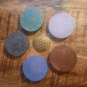 May include: Six round coasters with a geometric pattern in various shades of blue, brown, and white. A gold metal charm with a flower of life design is in the center.