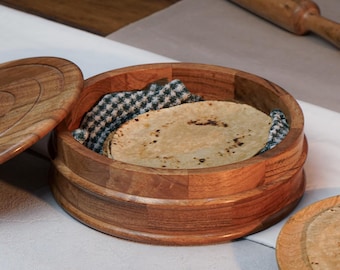 Puede incluir: Un recipiente redondo de madera con tapa a juego, que presenta un acabado de veta de madera natural. En el interior, un paño de tela a cuadros recubre el fondo, conteniendo varios alimentos planos, redondos y claros apilados.