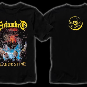 Puede incluir: Camiseta negra con el nombre de la banda "Entombed" en amarillo y rojo, y el título del álbum "Clandestine". La parte trasera de la camiseta muestra un diseño de calavera amarilla dentro de una luna creciente. Prenda de temática musical.
