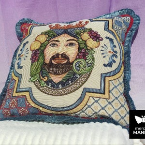 May include: A decorative pillow with a colorful tapestry design featuring a man's face with a crown and fruit. The pillow has a blue and red fringe border.